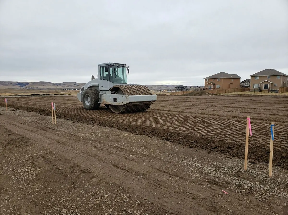 A padfoot roller with a studded steel drum compacts a layer of brown clay subgrade across a wide prepared area.